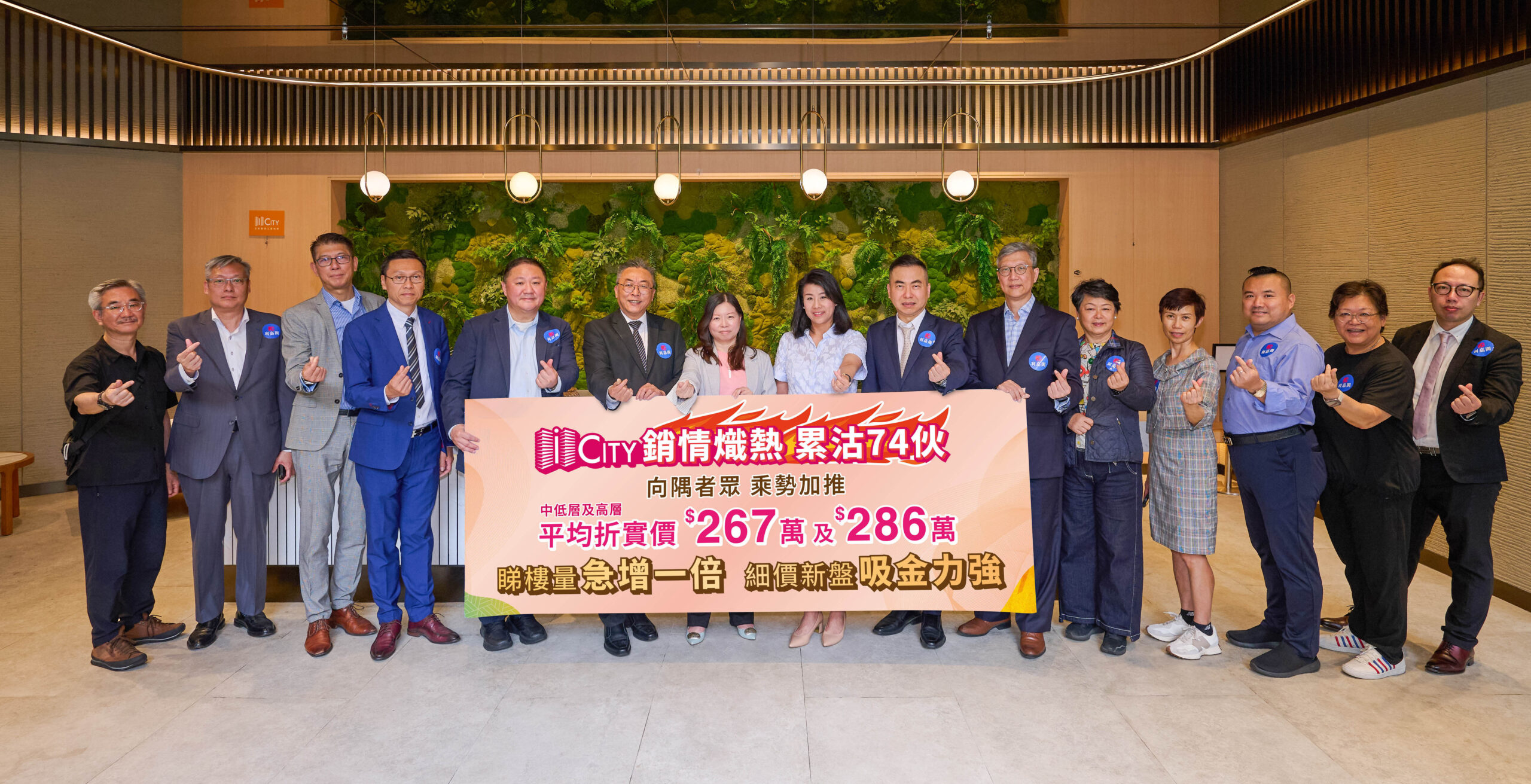 Group of professionals in business attire pose with a large pink banner in a modern lobby backdrop with green wall art behind them, signaling a promotional event.