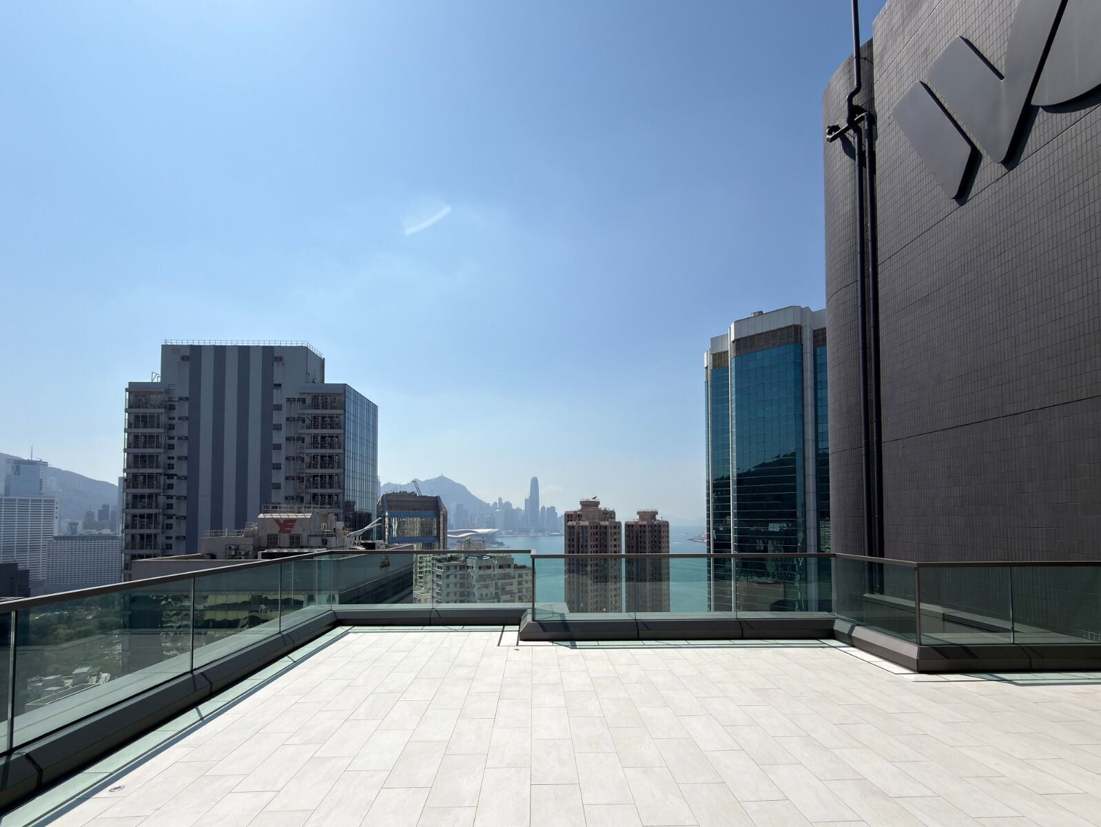 Rooftop terrace with glass railing overlooking a city skyline and harbor on a sunny day.