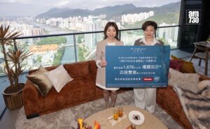 Two women in beige outfits hold a large blue promotional board inside a high rise living room with a panoramic city skyline and river behind them AM730 logo appears in the top right corner