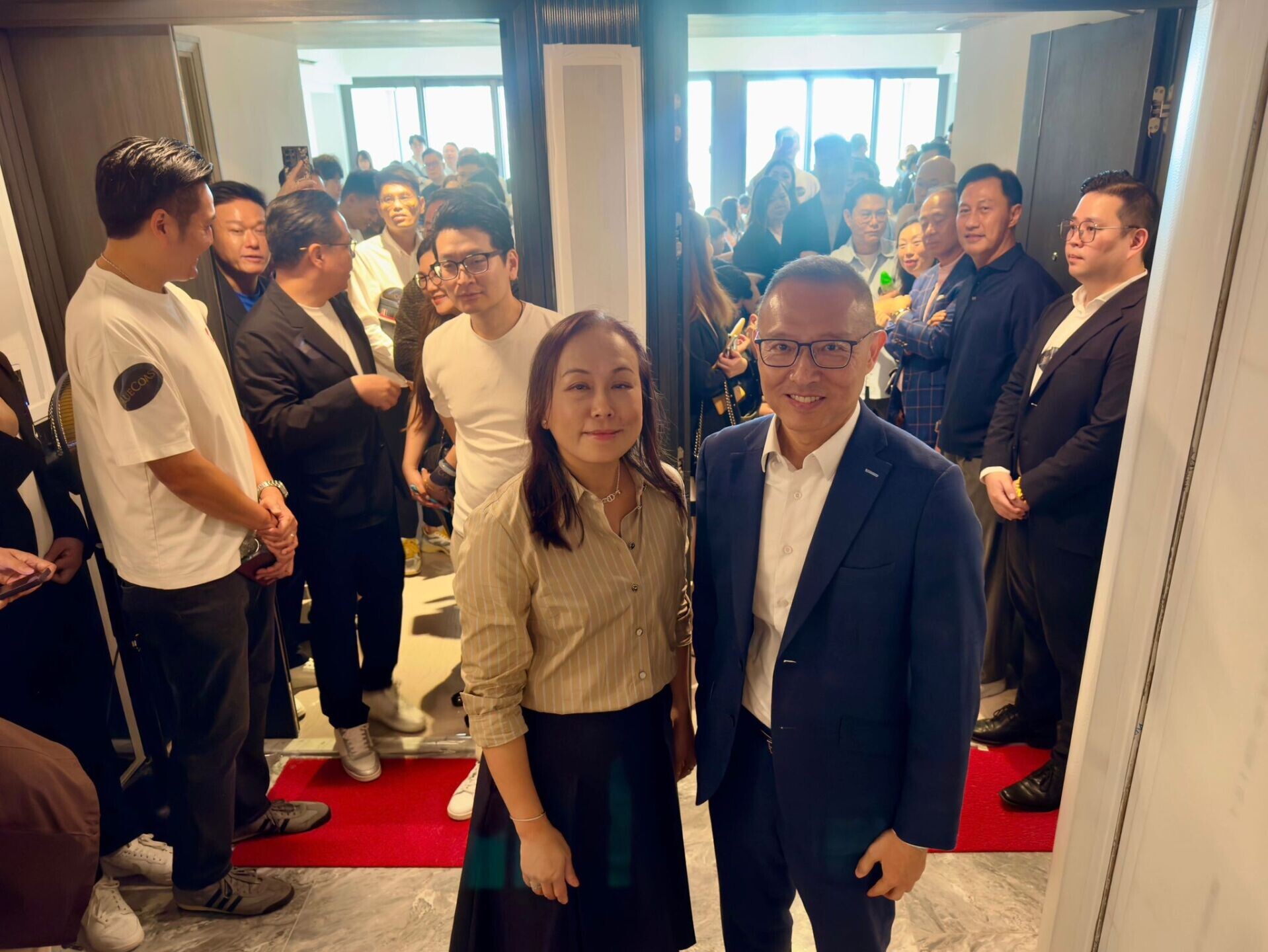 Two people in the foreground pose for a photo as a crowd of guests gathers in a hallway with open elevator doors behind them.