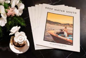 Stack of magazines titled Deep Water South cover shows a couple in a wooden boat on sunset water with flowers on a marble surface nearby
