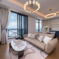 Bright open living room with beige sectional sofa white pillows and a glass top coffee table facing large windows and balcony doors Italianate chandelier overhead