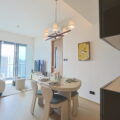 Bright open plan living and dining area with a round dining table set for six beige chairs and a modern chandelier plus large windows showing a city skyline