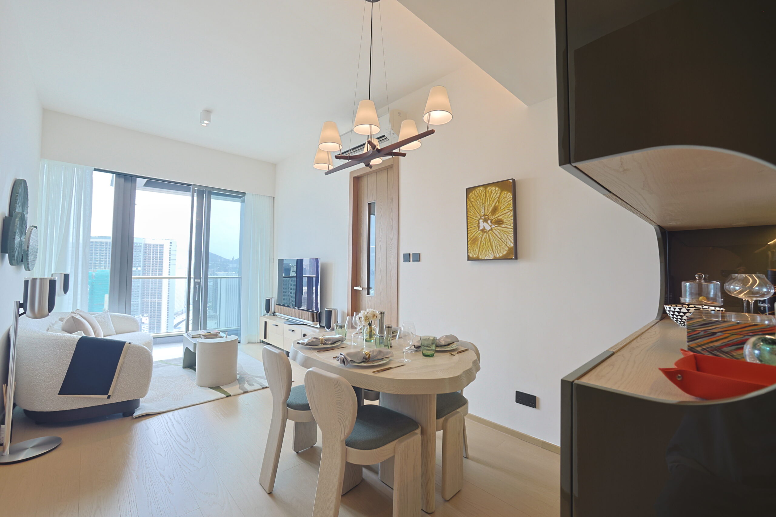 Bright open-plan living and dining area with a round dining table set for six, beige chairs, and a modern chandelier, plus large windows showing a city skyline.