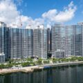 Row of tall glass apartment towers along a waterfront with construction cranes on top and a tree lined promenade in front