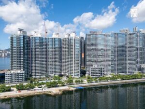 Row of modern glass apartment towers along a river with trees and a promenade in front and blue sky