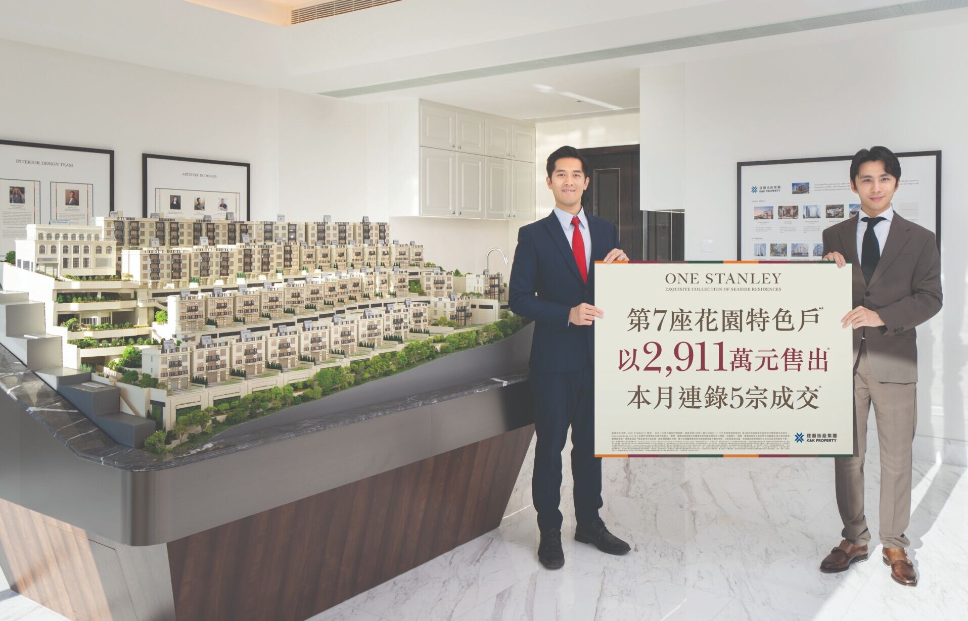 Two men in business suits pose in a real estate showroom, holding a large sign beside a detailed architectural model of a residential development.