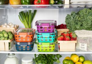 Open fridge scene showing colorful plastic baskets filled with assorted produce on a shelf including kale berries and citrus nearby