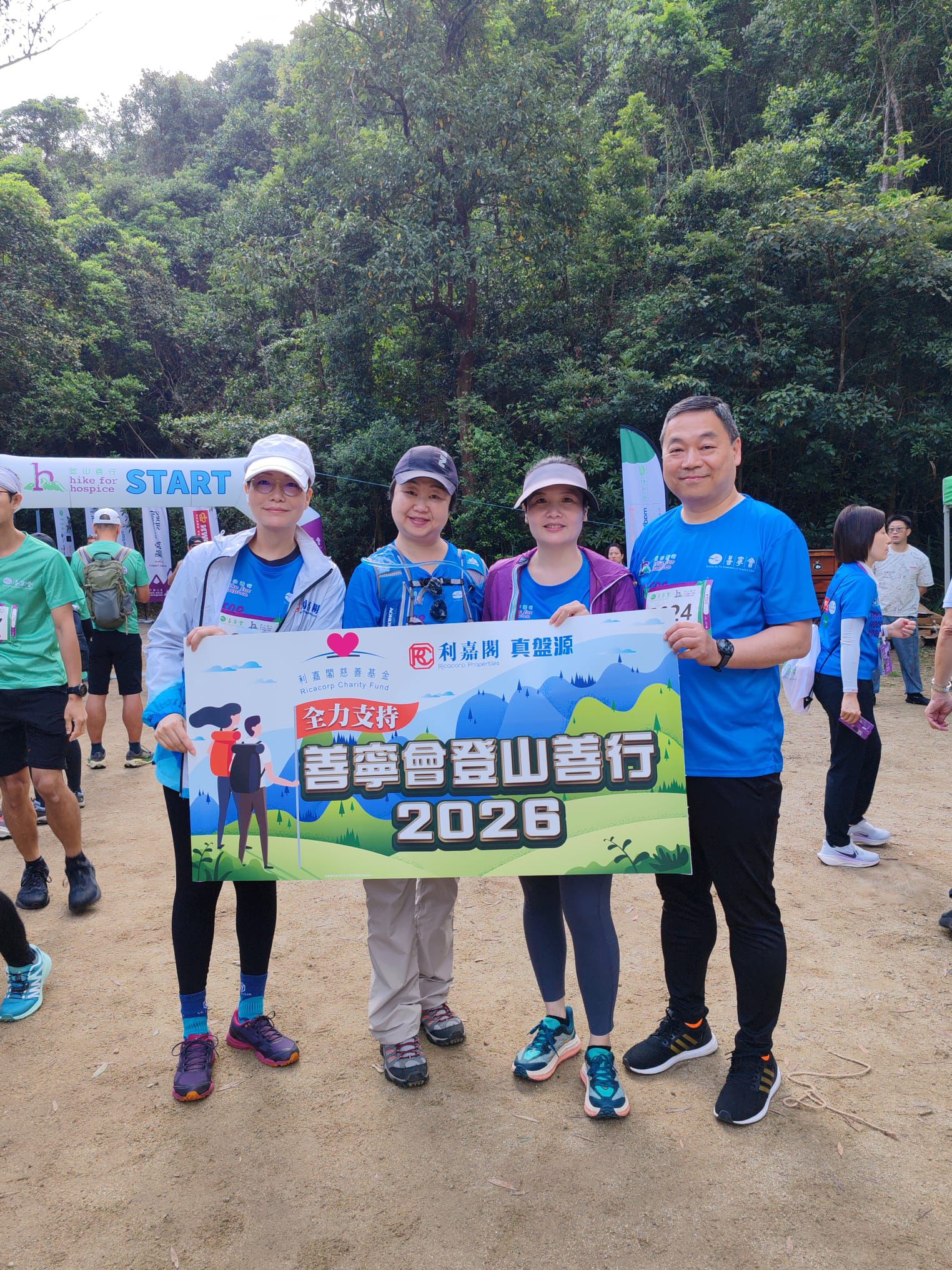 Four runners at the start line of a charity hike, holding a colorful banner with 2026 against a forest backdrop.