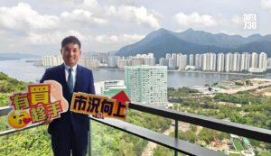 Man in a navy suit standing on a balcony holding colorful signs with Chinese characters overlooking a river high rise cityscape and mountains a logo appears in the top right corner
