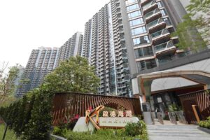 Tall modern high rise apartment complex with many curved balconies and glass facades near a gated entrance greenery in the foreground and a sign reading MARINA at the entrance