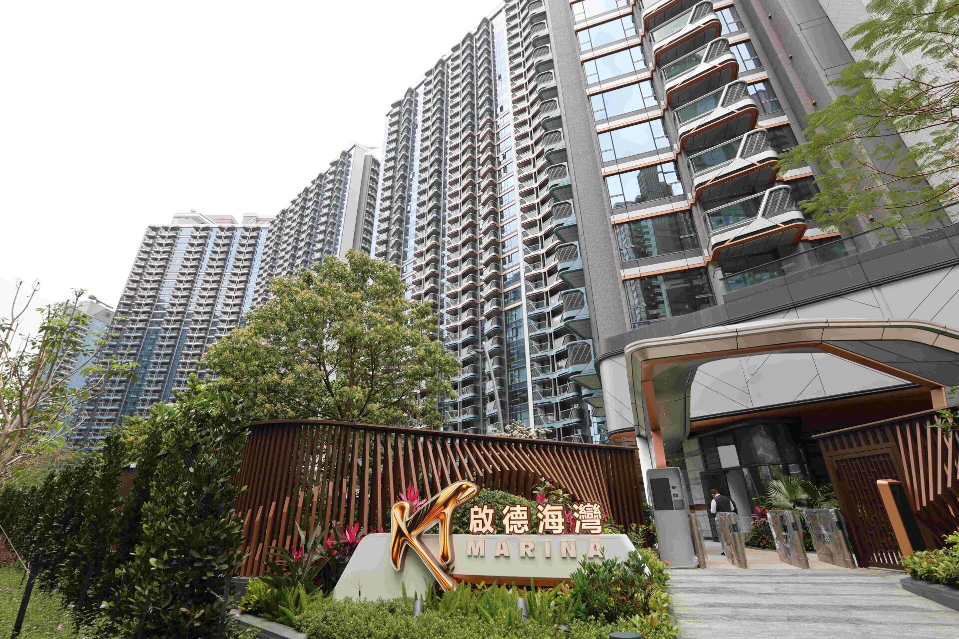 Tall modern high-rise apartment complex with many curved balconies and glass facades near a gated entrance, greenery in the foreground, and a sign reading “MARINA” at the entrance.
