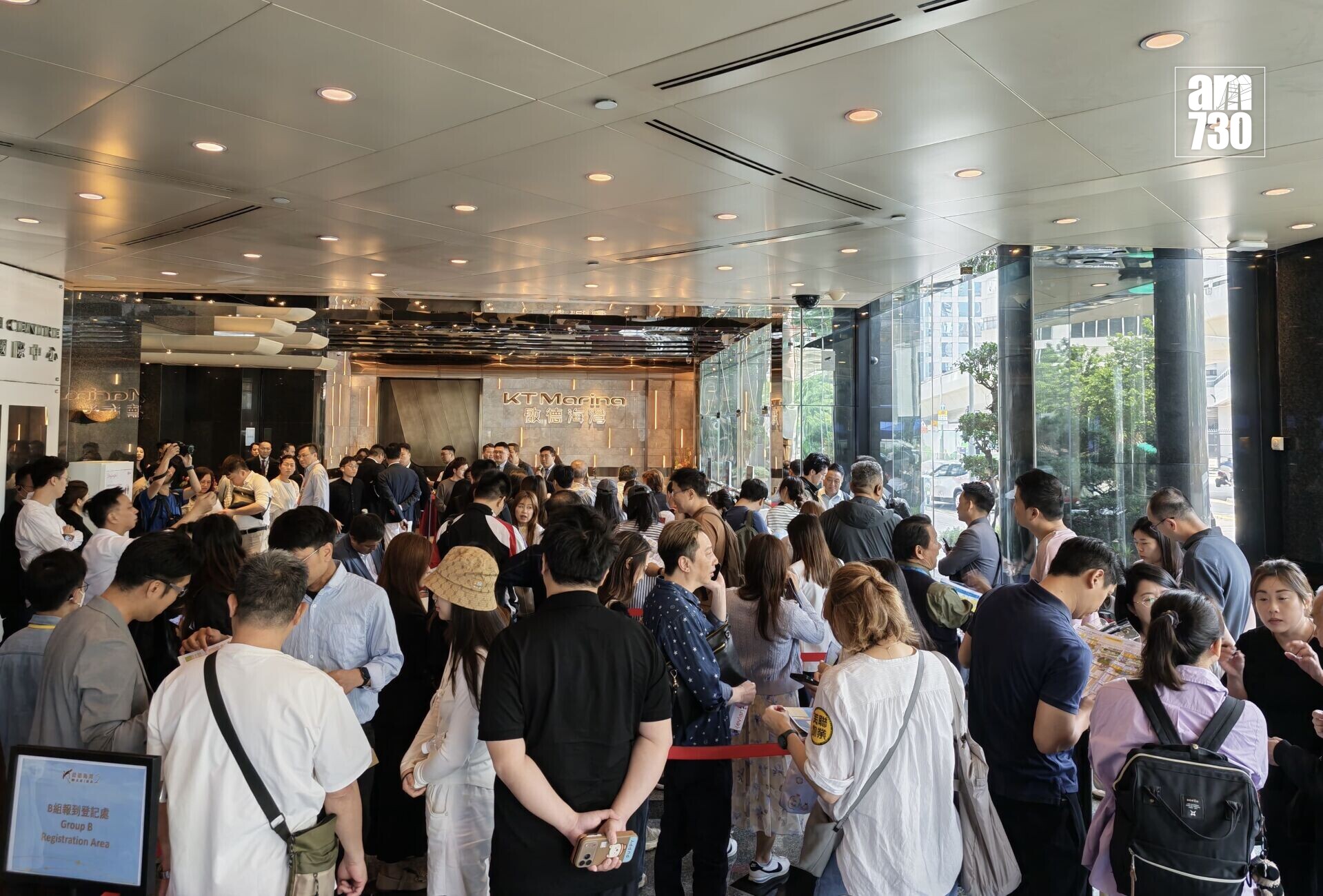 Crowded hotel lobby with people lining up near a reception desk, glass walls and bright ceiling lights.