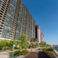 Row of glass residential towers along a waterfront promenade under a clear blue sky AM730 logo in the top right