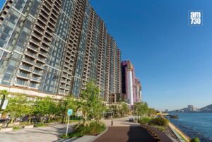 Row of glass residential towers along a waterfront promenade under a clear blue sky AM730 logo in the top right