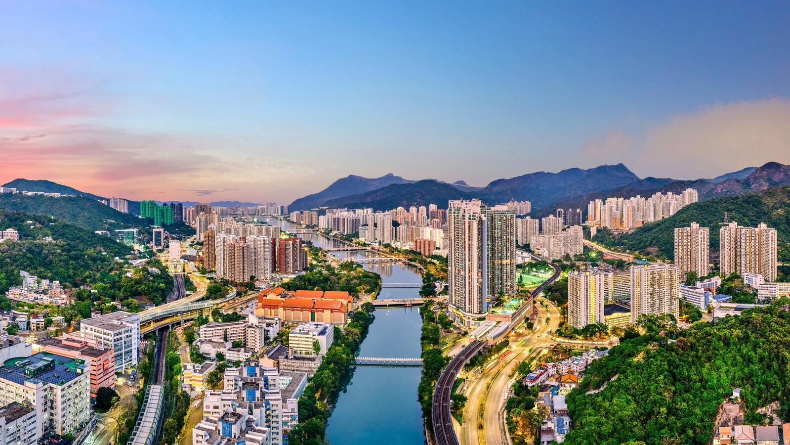 A dense cityscape with high-rise buildings along a winding river and surrounding green hills at sunset.