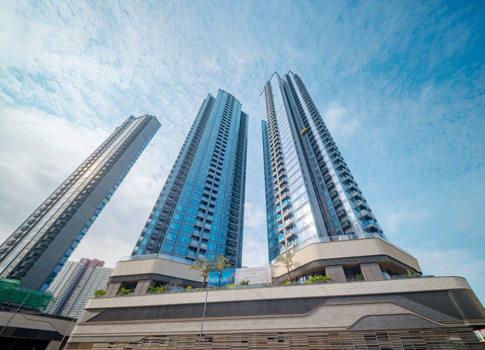 Three blue glass skyscrapers rise above a multi-level podium against a clear blue sky with palms at the base.