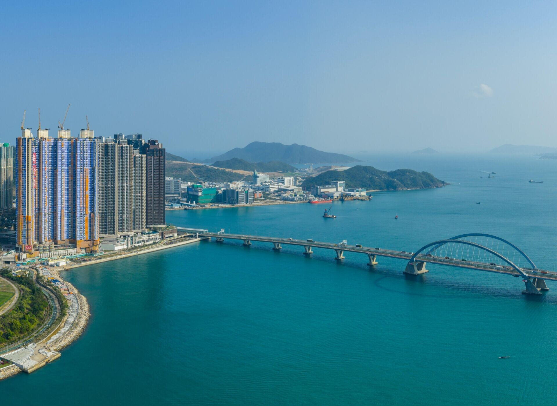 City skyline with high-rise buildings under construction along a harbor, a bridge crossing turquoise water.