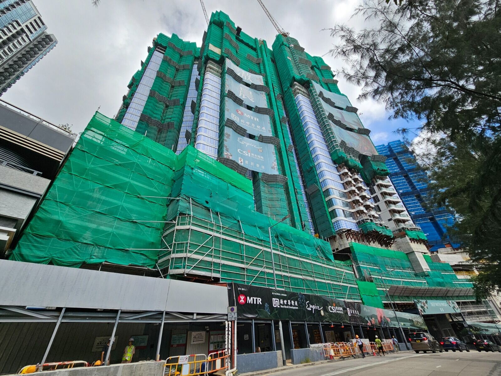 Under-construction skyscraper wrapped in green safety netting with scaffolding and a street-level construction barricade.