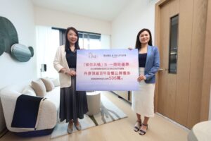 Two women in business attire stand indoors holding a large promotional banner with text and logos Bang  Olufsen in a bright living room setting