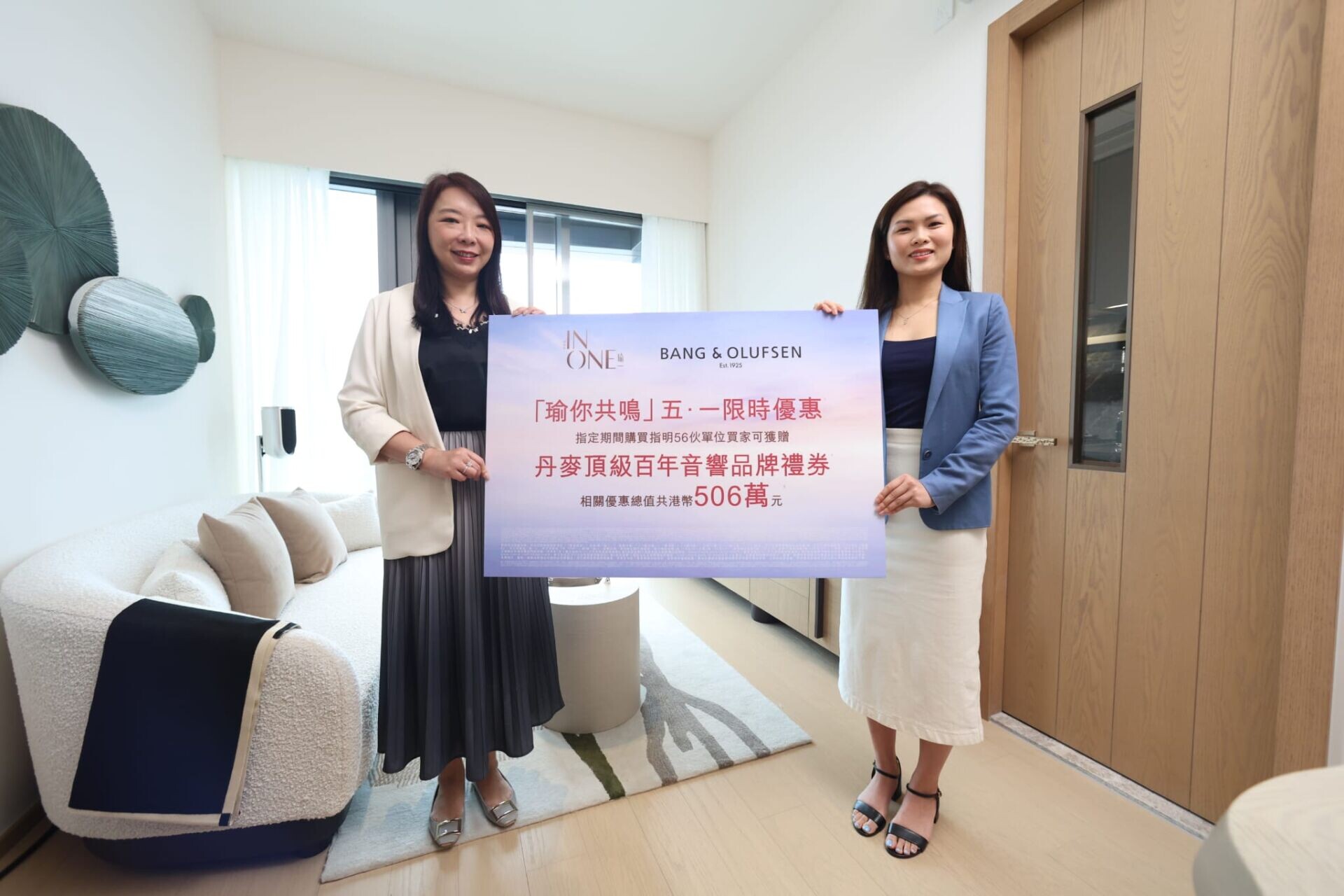 Two women in business attire stand indoors, holding a large promotional banner with text and logos (Bang & Olufsen) in a bright living room setting.