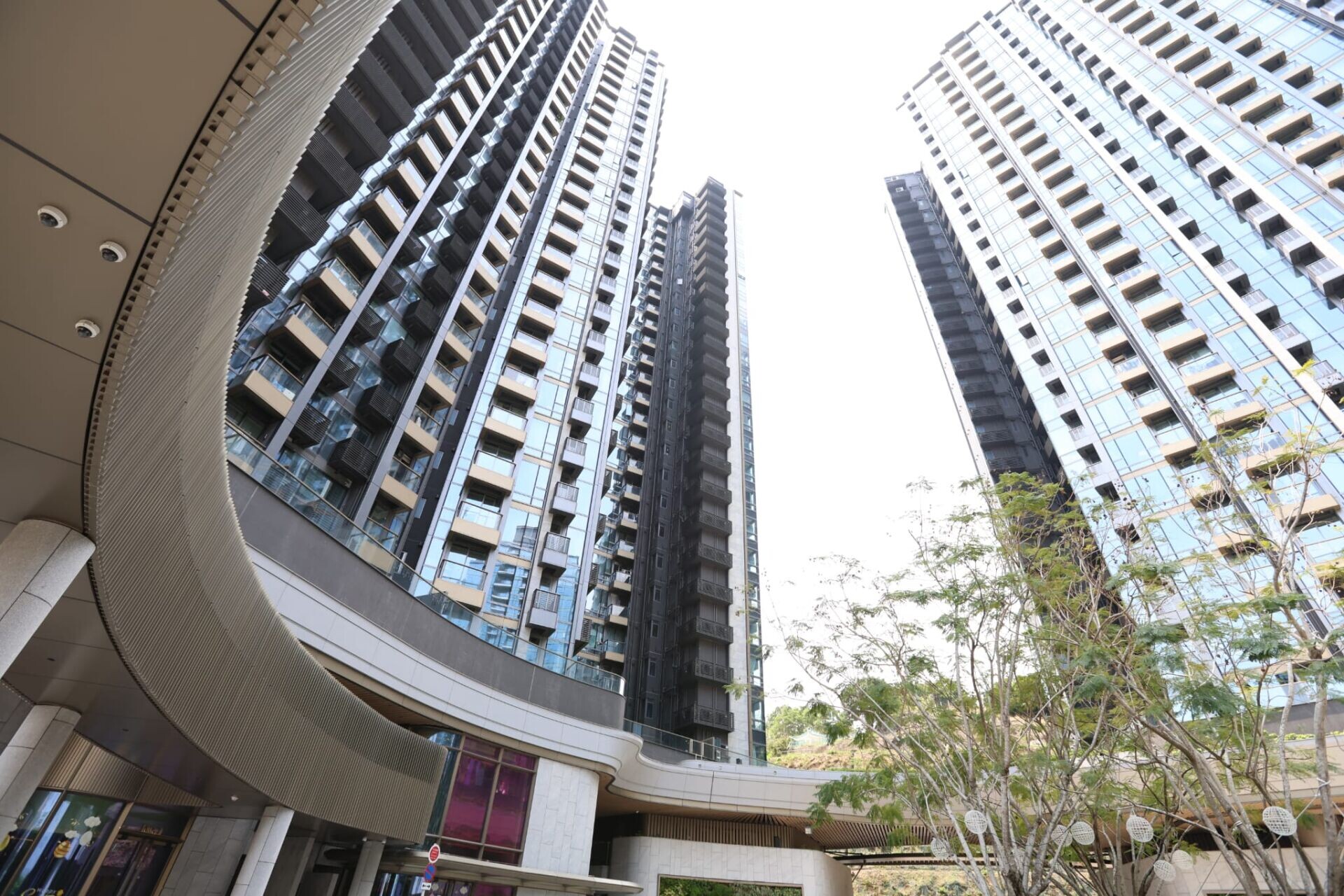 Low-angle view of tall glass office towers surrounding a curved commercial building, with trees in foreground.