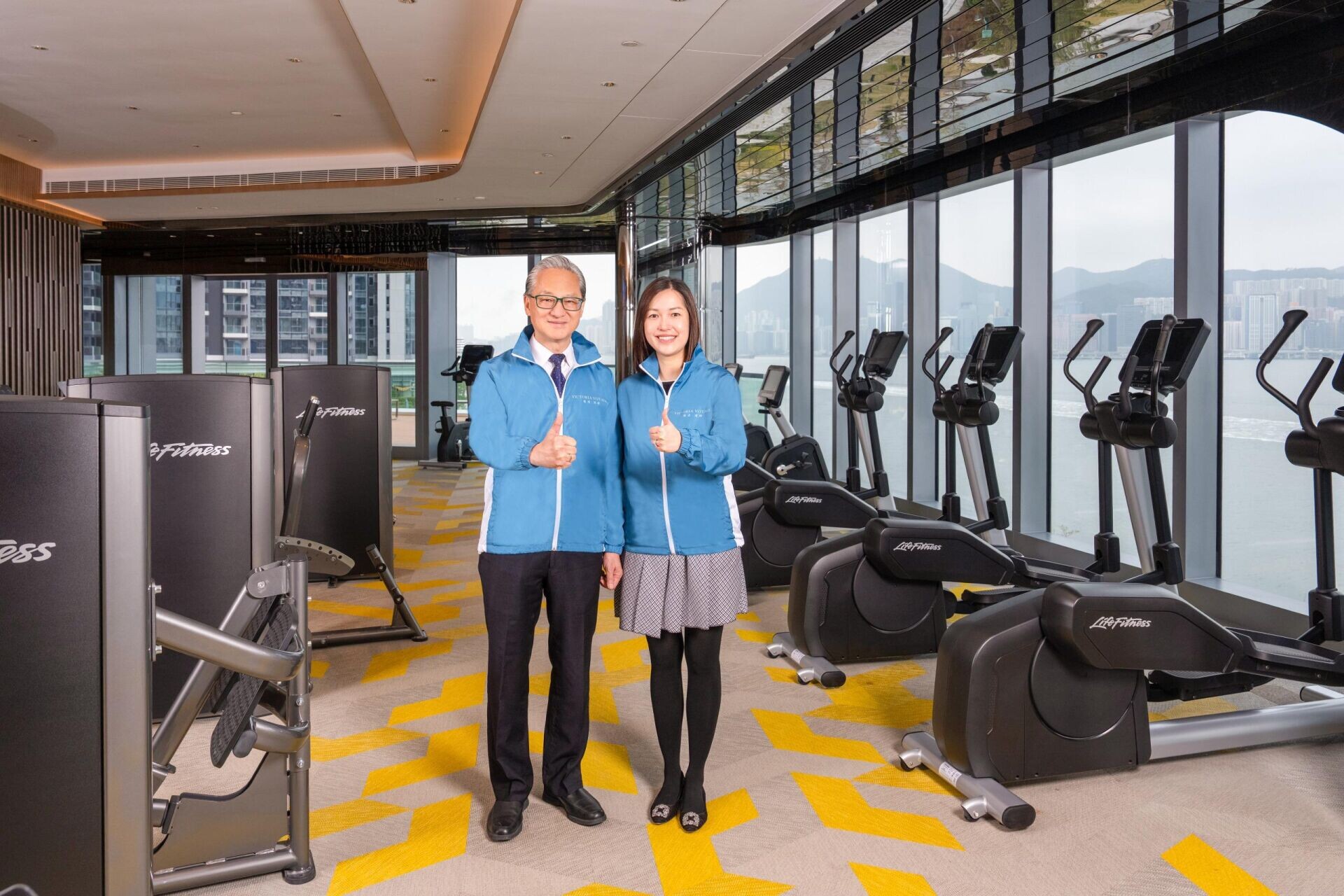 Two staff members in blue jackets posing and giving thumbs up in a bright gym with treadmills by large windows.