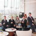 Five professionals in suits sit on a blue sofa in a bright lobby giving thumbs up with a city skyline seen through large glass panels and an AM730 logo in the corner