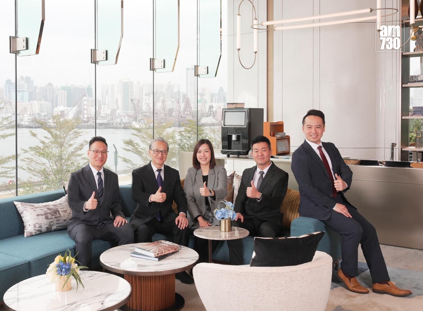 Five professionals in suits sit on a blue sofa in a bright lobby, giving thumbs up with a city skyline seen through large glass panels and an AM730 logo in the corner.