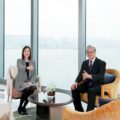 Two professionals a woman and a man sit in a bright lounge by large windows with a harbor view giving thumbs up