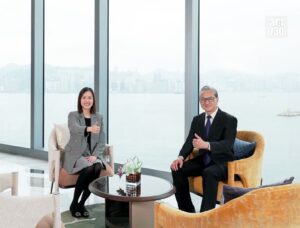 Two professionals a woman and a man sit in a bright lounge by large windows with a harbor view giving thumbs up