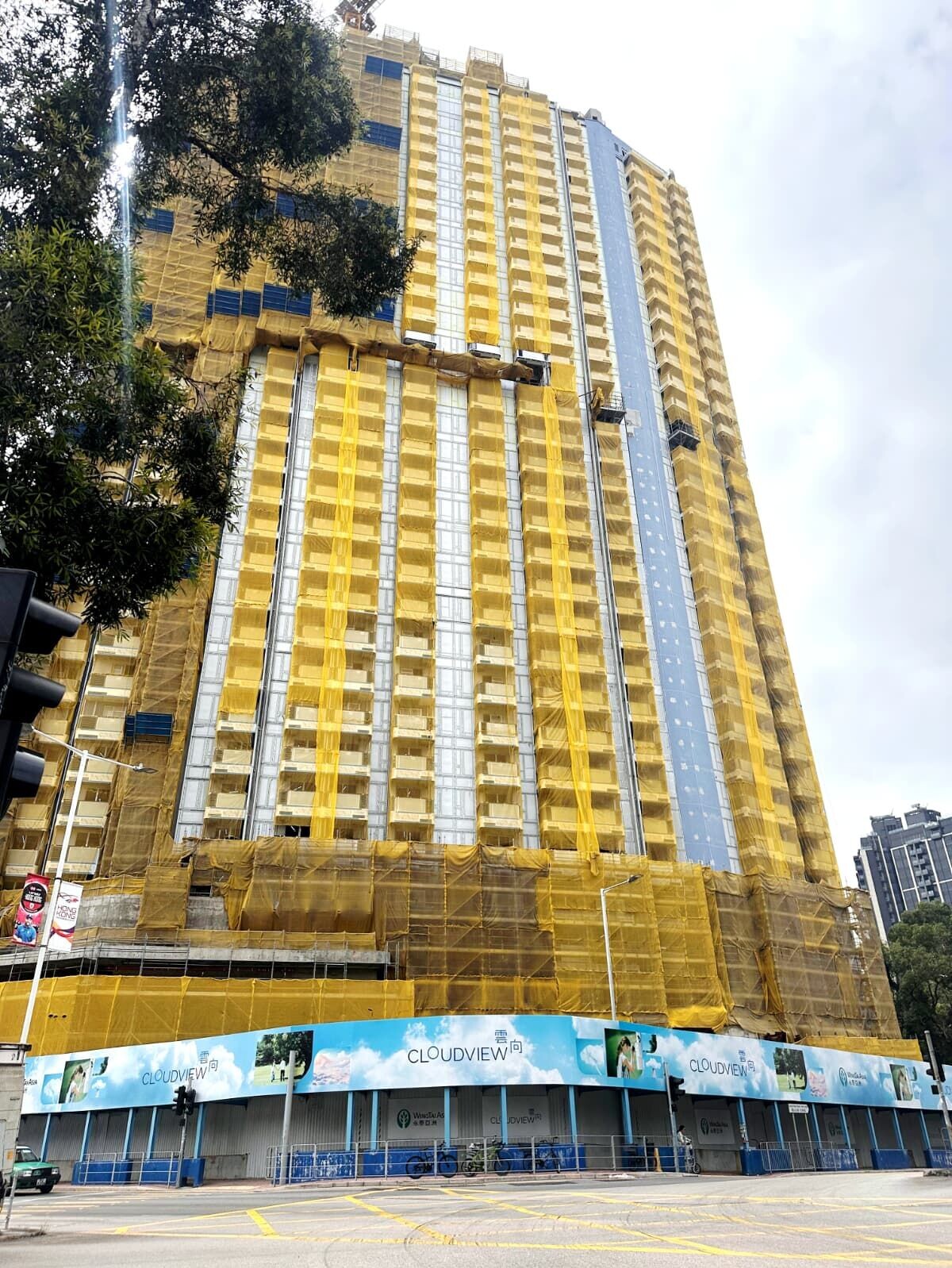 High-rise building under construction with yellow safety nets and blue glass panels, street level with construction barriers and banners reading CLOUDVIEW
