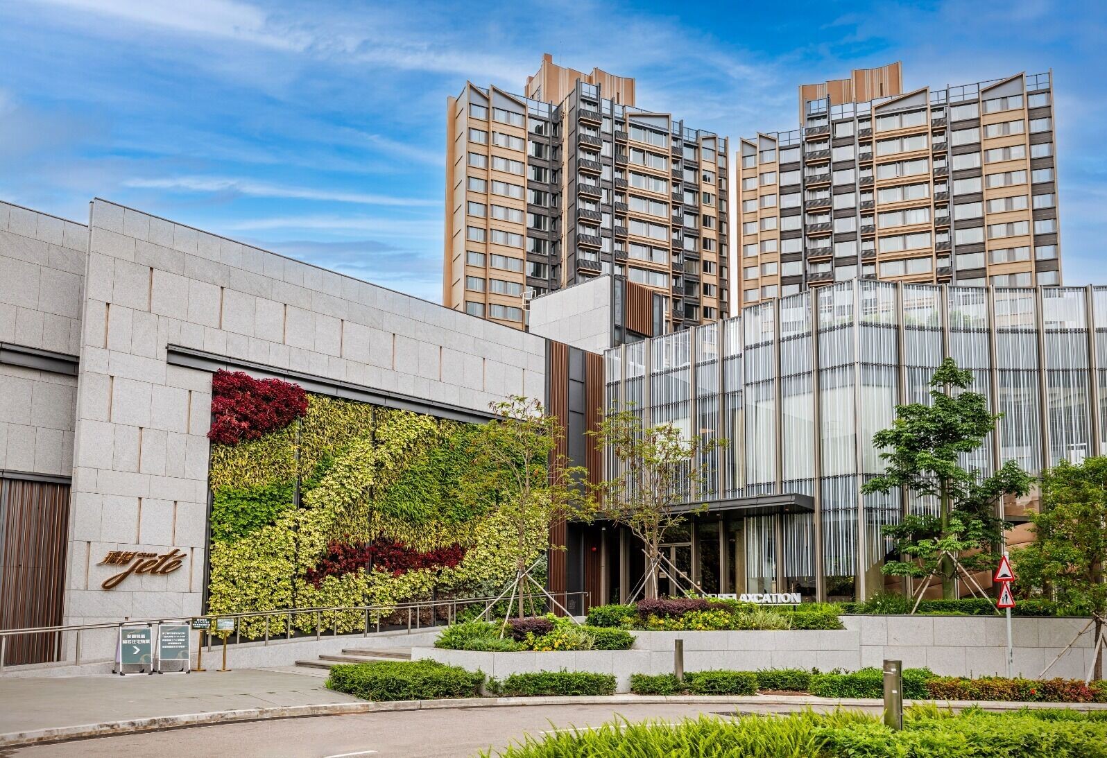 Modern urban complex with a vertical garden on a stone wall and glass facade, tall residential towers in the background against a blue sky.
