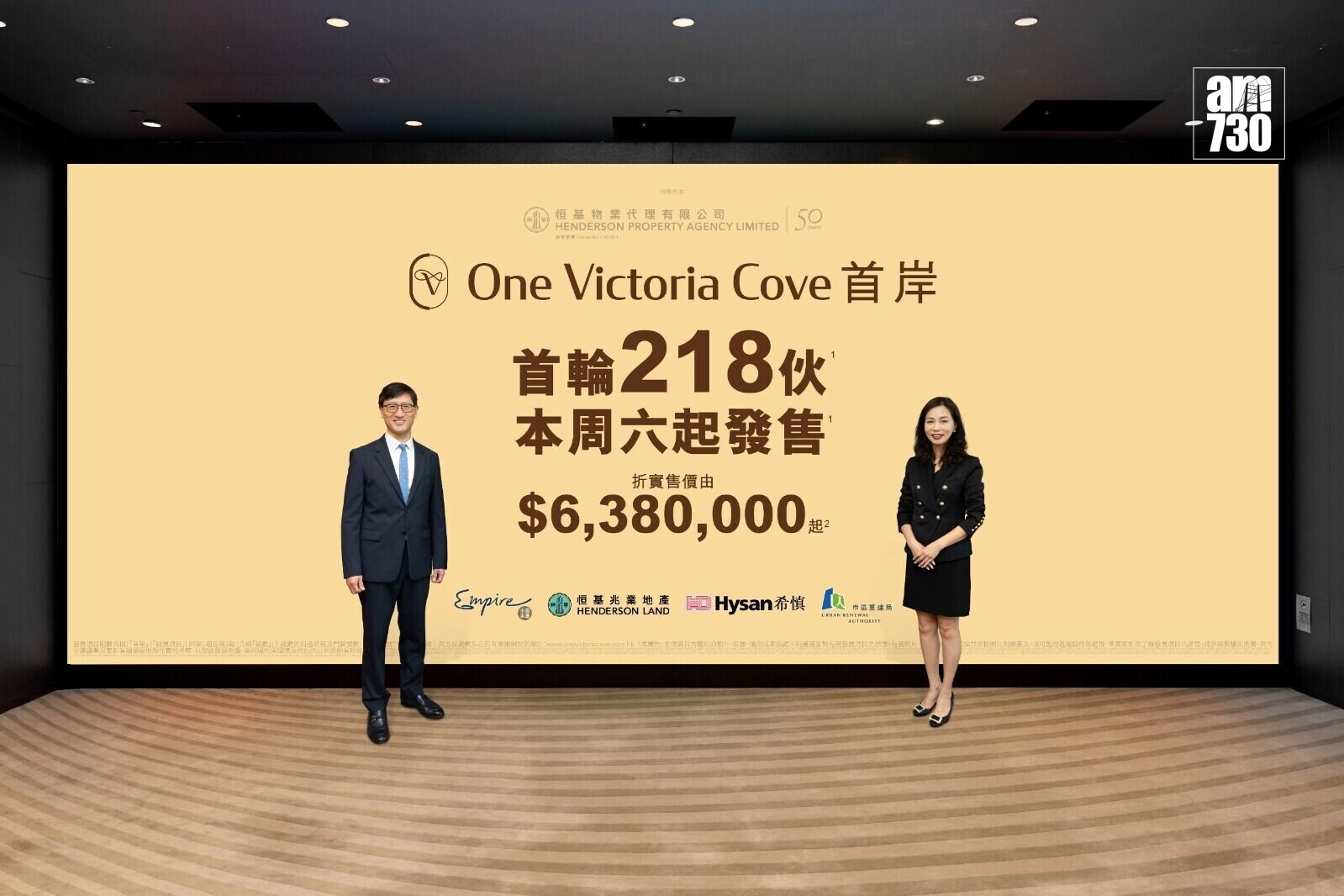 Two professionally dressed presenters, a man in a suit and a woman in a black outfit, stand beside a large beige promotional banner for ‘One Victoria Cove’ inside a showroom with a patterned carpet and dark walls.