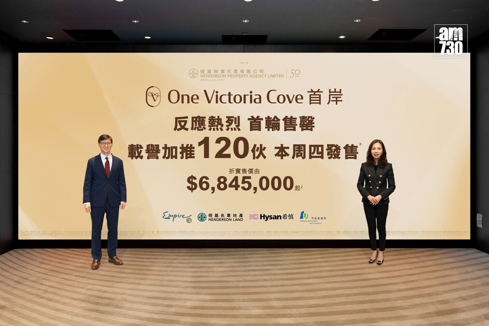Two business professionals stand on either side of a large beige promotional banner for One Victoria Cove.