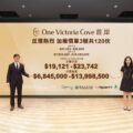 Two professionally dressed presenters stand on each side of a large beige promotional backdrop with bold pricing text and logos likely at a real estate or launch event