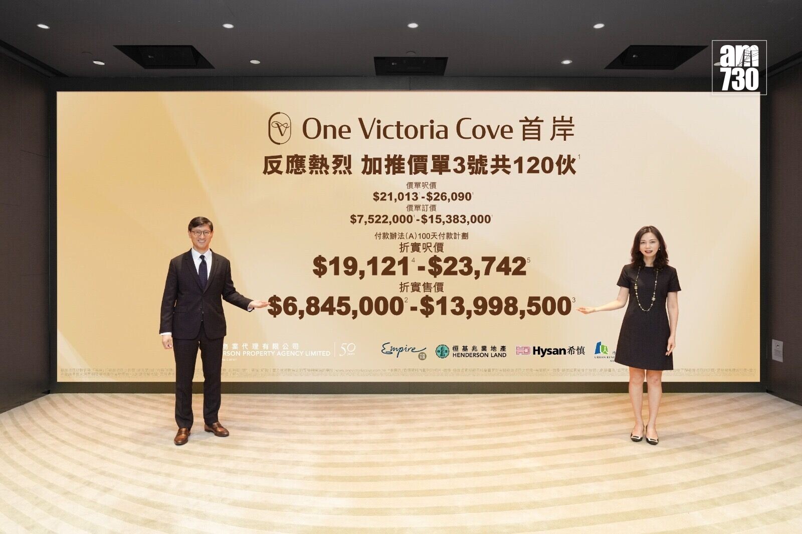 Two professionally dressed presenters stand on each side of a large beige promotional backdrop with bold pricing text and logos, likely at a real estate or launch event.