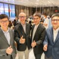 Four men in suits posing and giving thumbs up at a bustling indoor event with glass walls and tables in the background