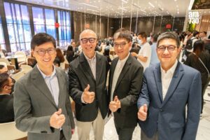 Four men in suits posing and giving thumbs up at a bustling indoor event with glass walls and tables in the background