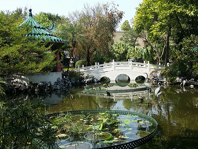 05 | 利嘉閣地產有限公司 Chinese garden scene with a white arched bridge over a pond and a teal roof pavilion among trees