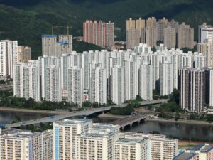 Dense row of white high rise apartment blocks beside a river with a bridge and green hills in the background