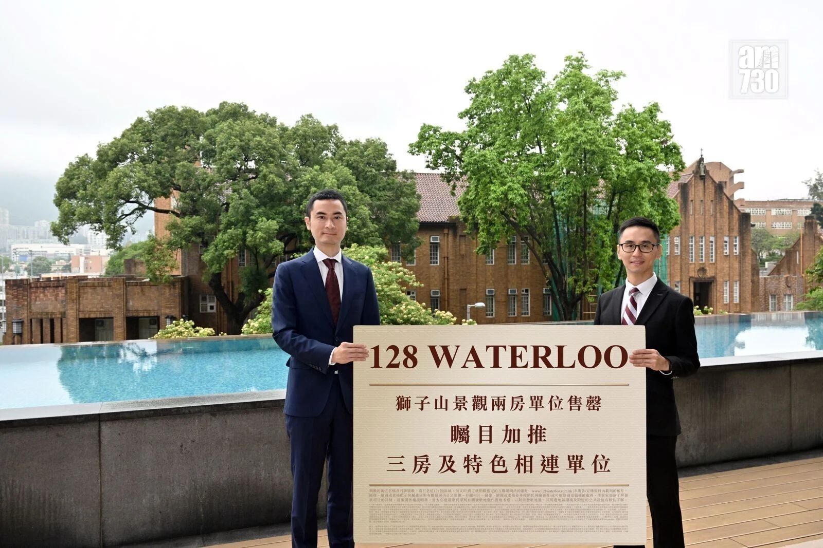 Two men in suits hold a large sign reading '128 WATERLOO' outdoors by a pool, with green trees and brick buildings in the background.