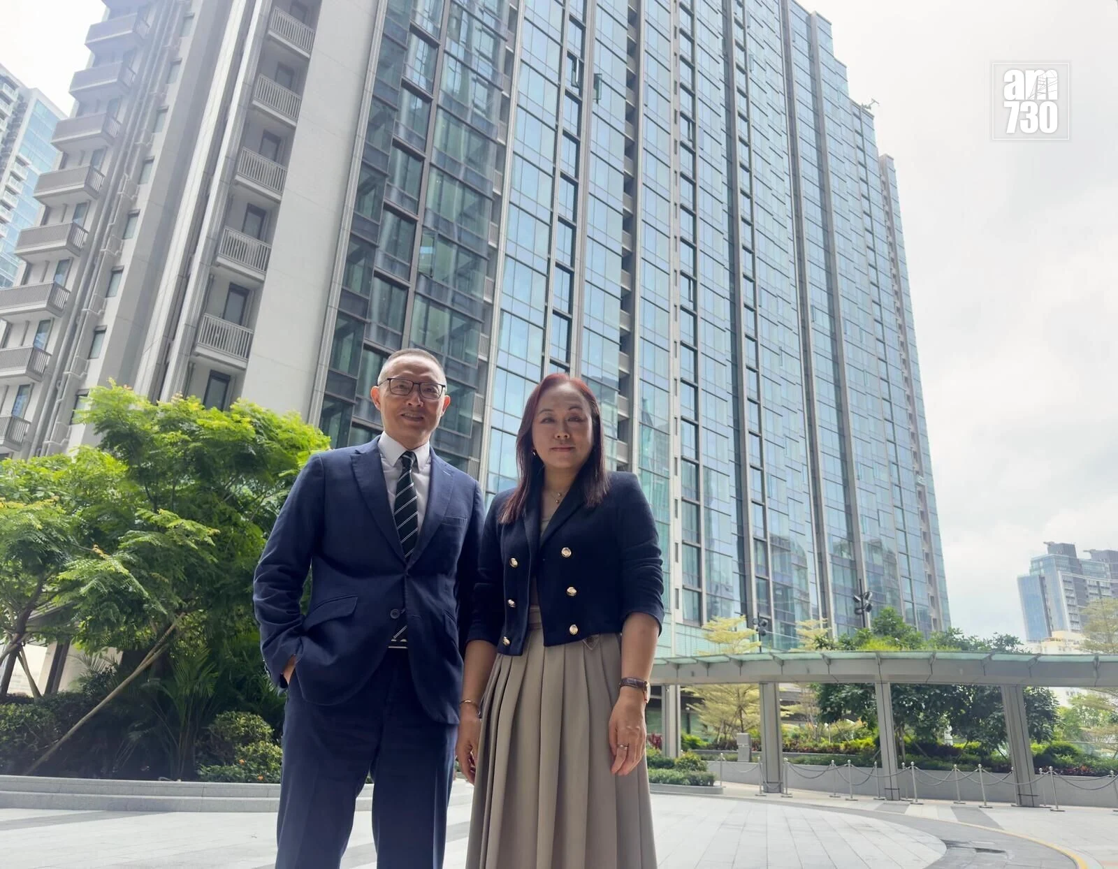 Two professionally dressed people pose outside a glass-fronted office building (man in a navy suit, woman in a blazer and beige skirt).