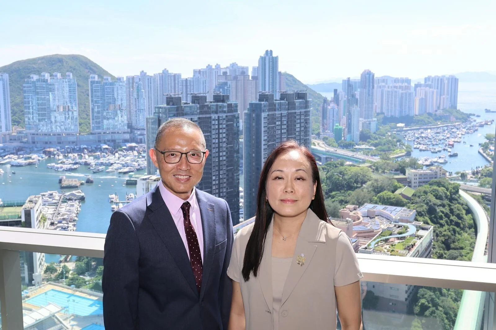 Two smiling professionals posing on a balcony with a city skyline and marina in the background.