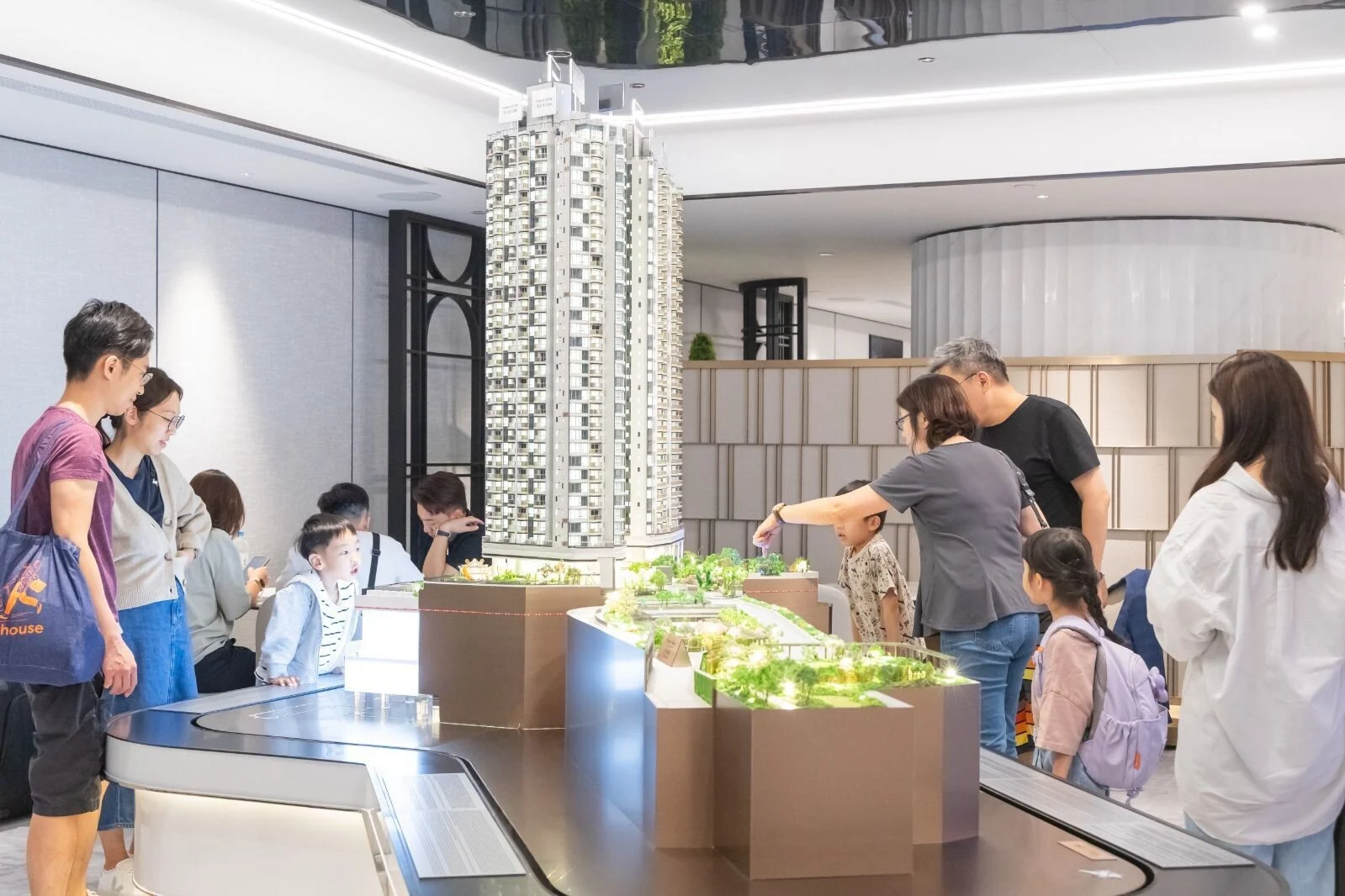 People gather around a large detailed scale model of a tall residential tower in a showroom or exhibition space, examining the model closely.