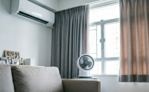 Cozy living room corner with a beige sofa wall AC framed photos on a shelf a white fan by a window and magazines on a light wood coffee table