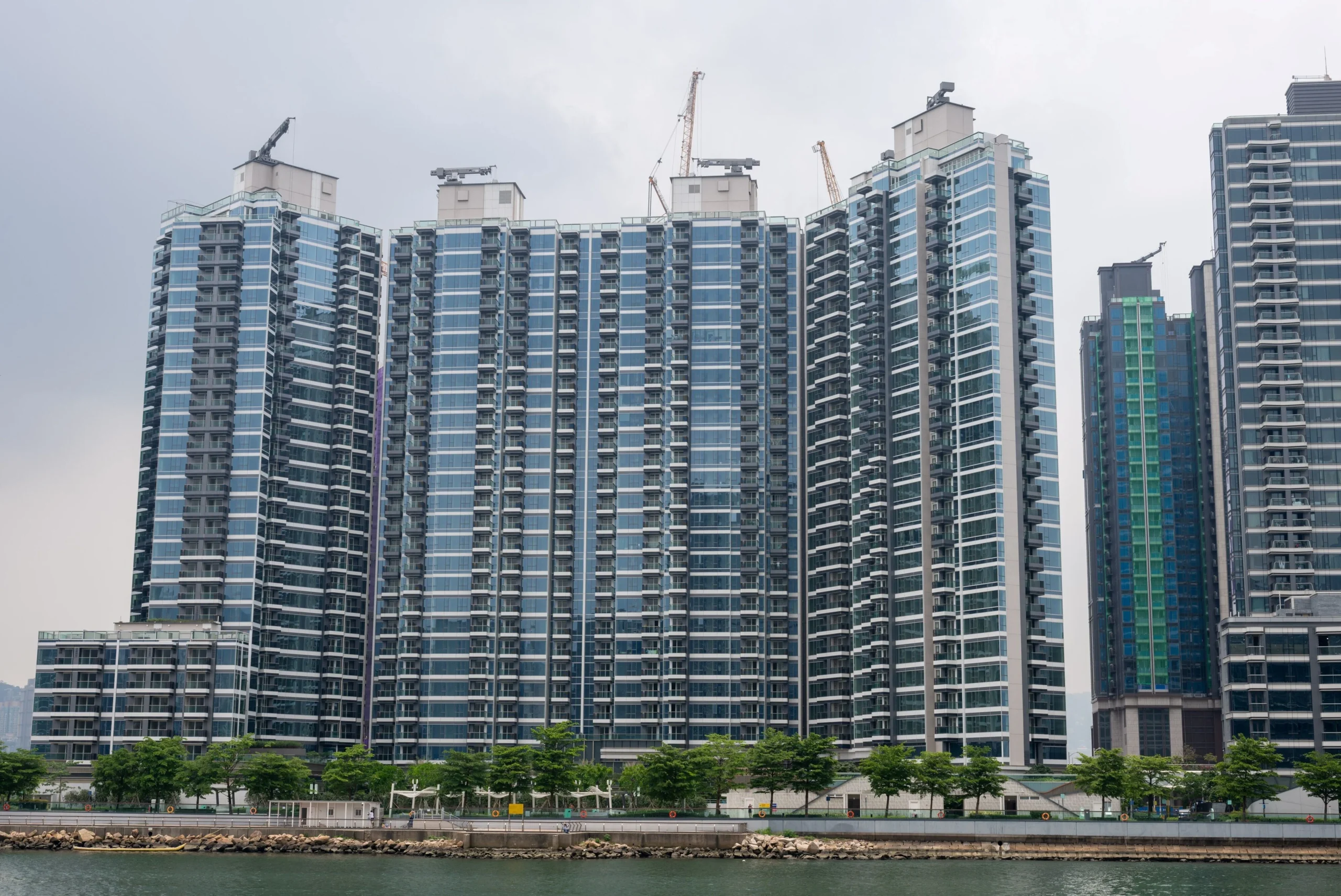 Row of blue glass high-rise apartment buildings along a waterfront promenade with construction cranes on the rooftops and trees in front.