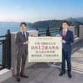 Two men in business suits stand on a seaside terrace holding a large sign with Chinese text ocean and mountains in the background