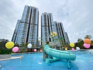 Tall glass residential towers with a turquoise waterslide in a pool area surrounded by colorful balloons and amenities