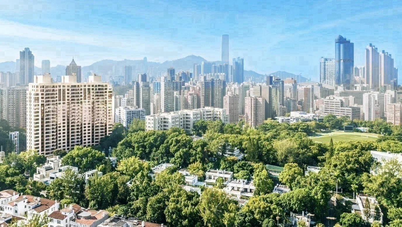 Aerial view of a dense city skyline with tall skyscrapers and a large green park in the foreground under a blue sky, mountains in the distance.